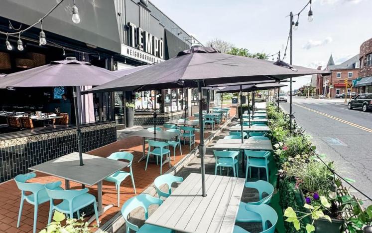 Tempo restaurant patio on Moody street adorned with various teal chairs and tables under umbrellas on a warm, sunny day.