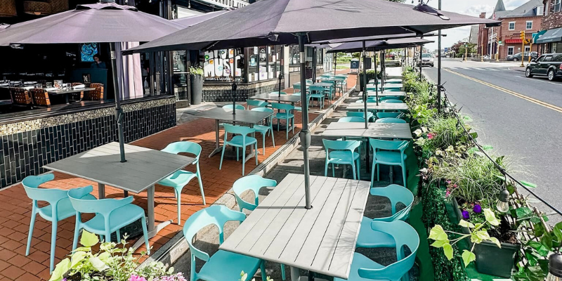Tempo restaurant patio on Moody street adorned with various teal chairs and tables under umbrellas on a warm, sunny day.